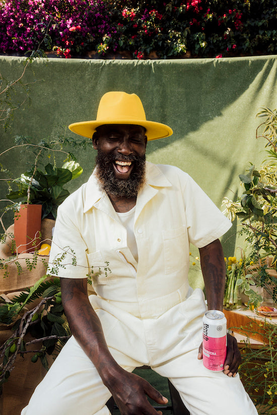 Hakim Tafari smiling wearing a white jump suit and yellow hat and holding a can of Better Booch