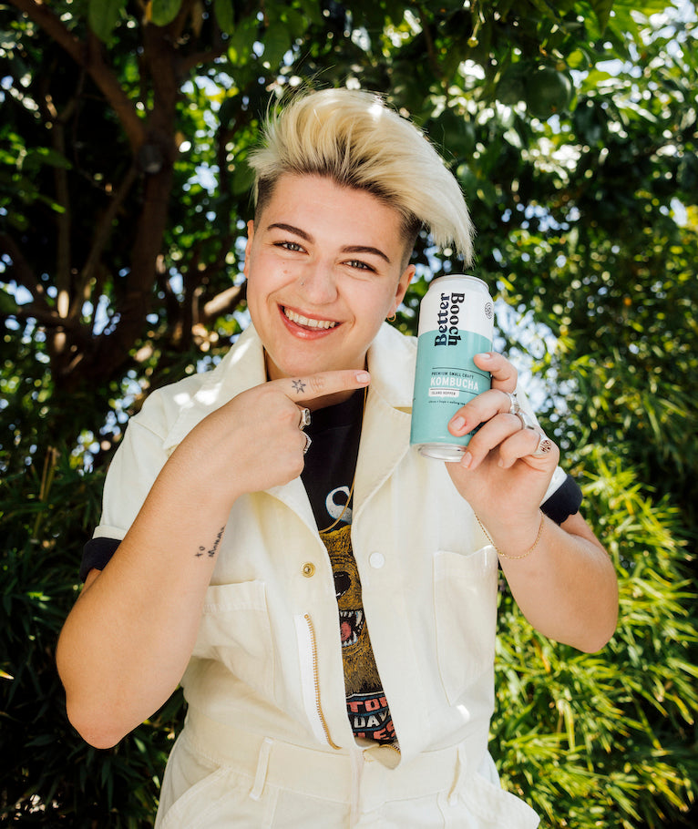 Artist September standing under a tree and pointing to a can of Better Booch organic kombucha